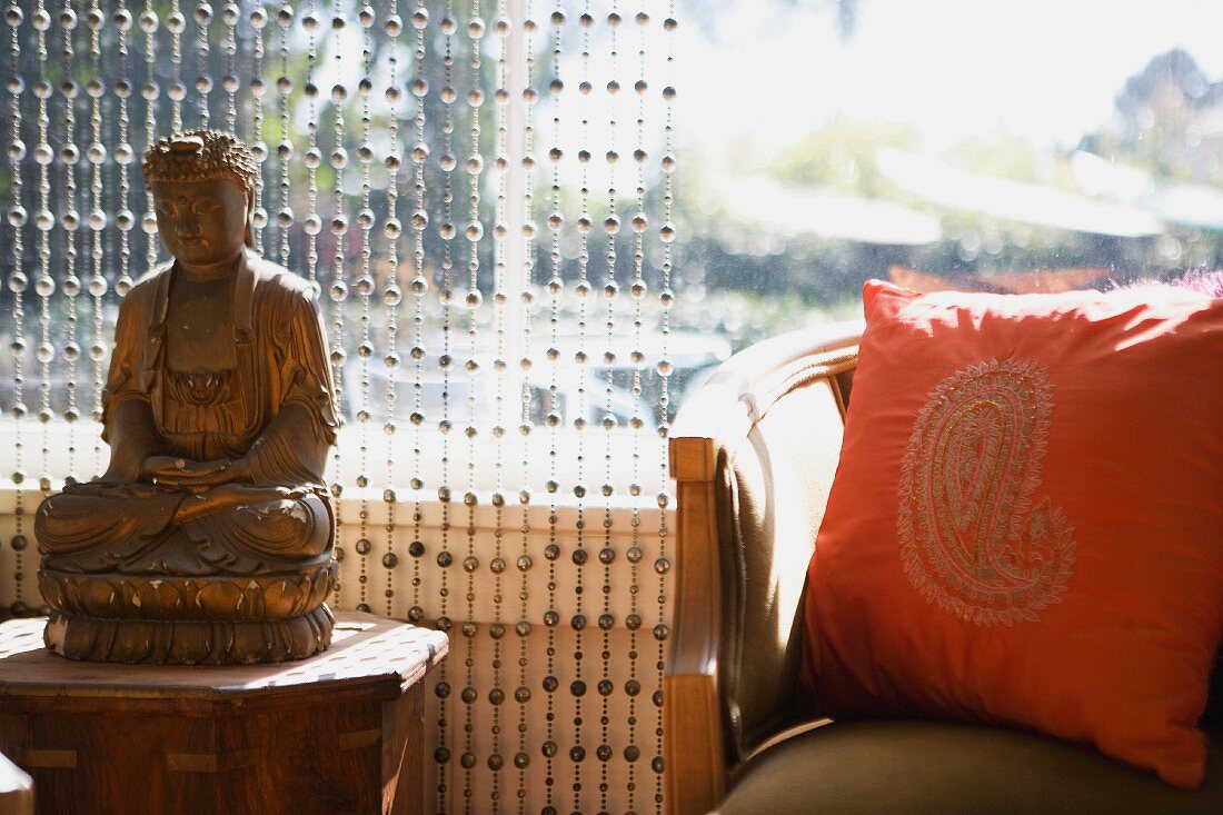 Buddhafigur aus Holz vor Vorhang mit aufgefädelten Kugeln und folkloristisches Kissen auf Armlehnstuhl