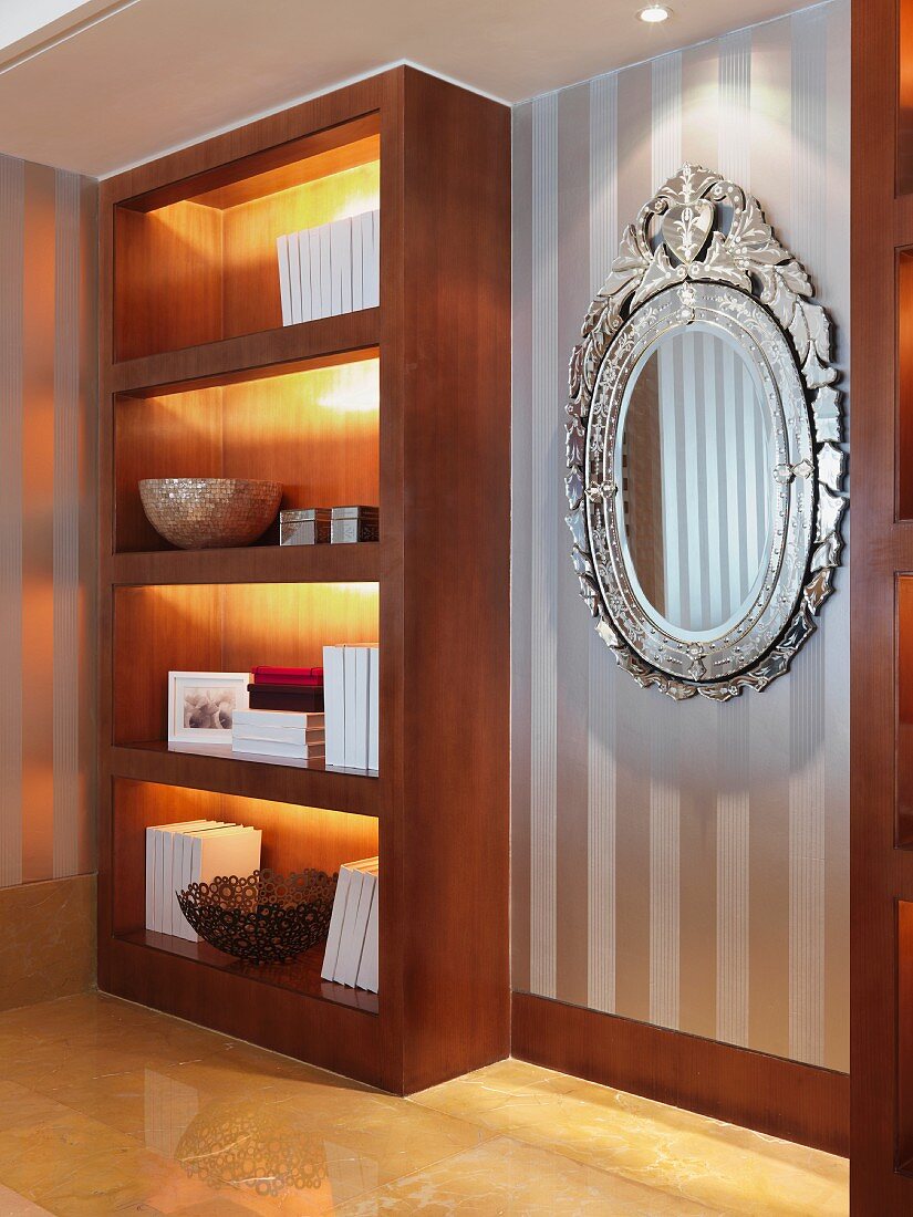 Illuminated, fitted shelving next to mirror with ornate silver frame on wall with striped wallpaper