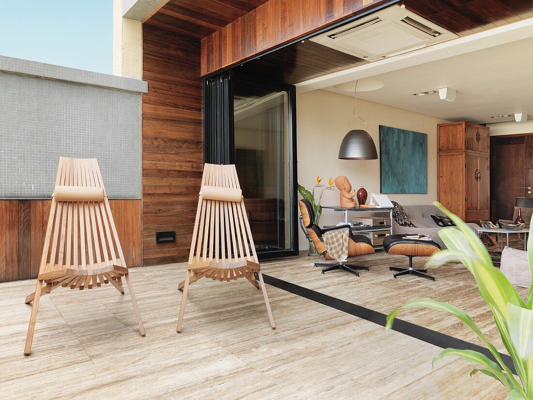 Outdoor chairs made of wooden slats on terrace with stone floor and view into open-plan interior