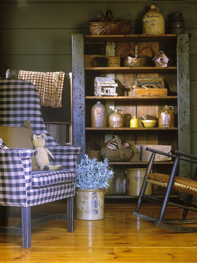 Holzregal im Vintagestil mit einer Antiquitätensammlung; daneben ein Polsterstuhl mit weiss-blauem Karomuster und ein Schaukelstuhl