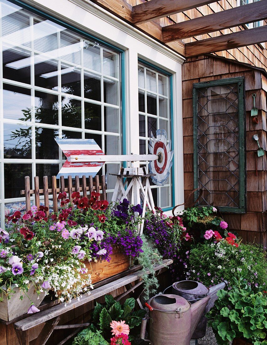 Fensterfront mit Windrad, einer einfachen Holzbank und darauf stehenden, blühenden Blumenkästen