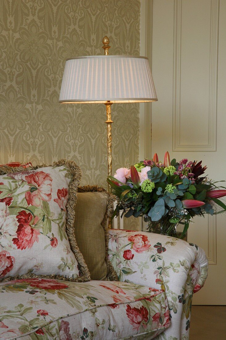 Geblümtes Sofa mit Kissen im gleichen Stoff neben Stehlampe und Blumenstrauss im Halbdunkel