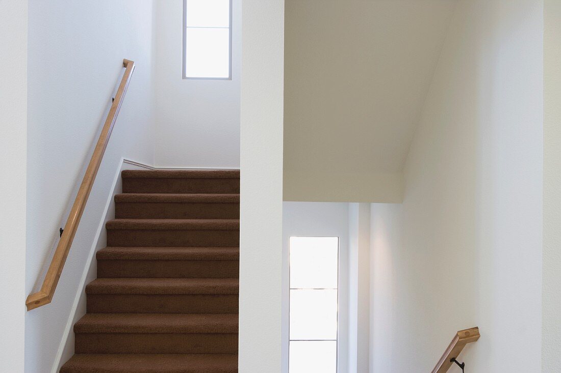 Weisses, schlichtes Treppenhaus mit braunem Teppichbelag auf Stufen und Handlauf aus Holz
