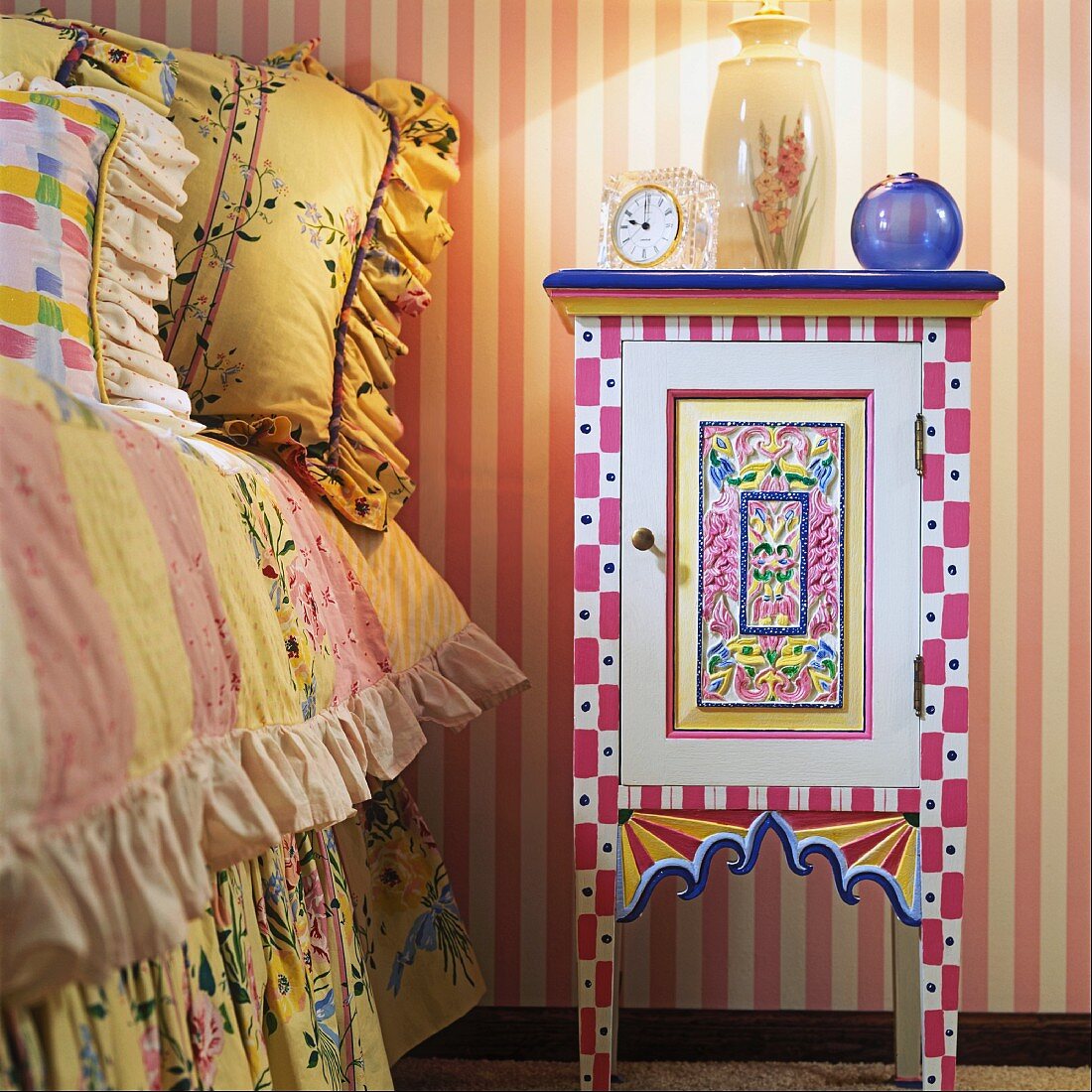 Selbstbemalter, farbenfroher Nachttisch neben Kinderbett mit verspielten Kissen und Bettdecke mit Rüschen; an der Wand eine rosa-weiss gestreifte Tapete