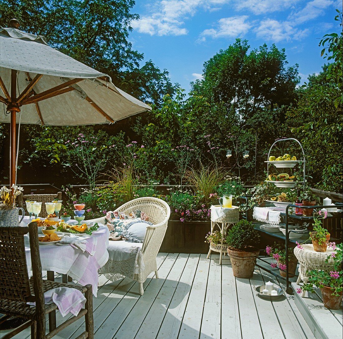 Wicker armchair and many potted plants on sunny wooden terrace with parasol and laid dining table