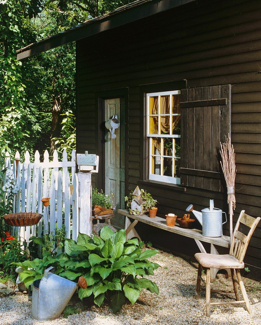 Einfaches Holzhaus im Grünen mit Schiebefenster, weißem Gartenzaun, Gartenutensilien und einer verwitterten Sitzbank
