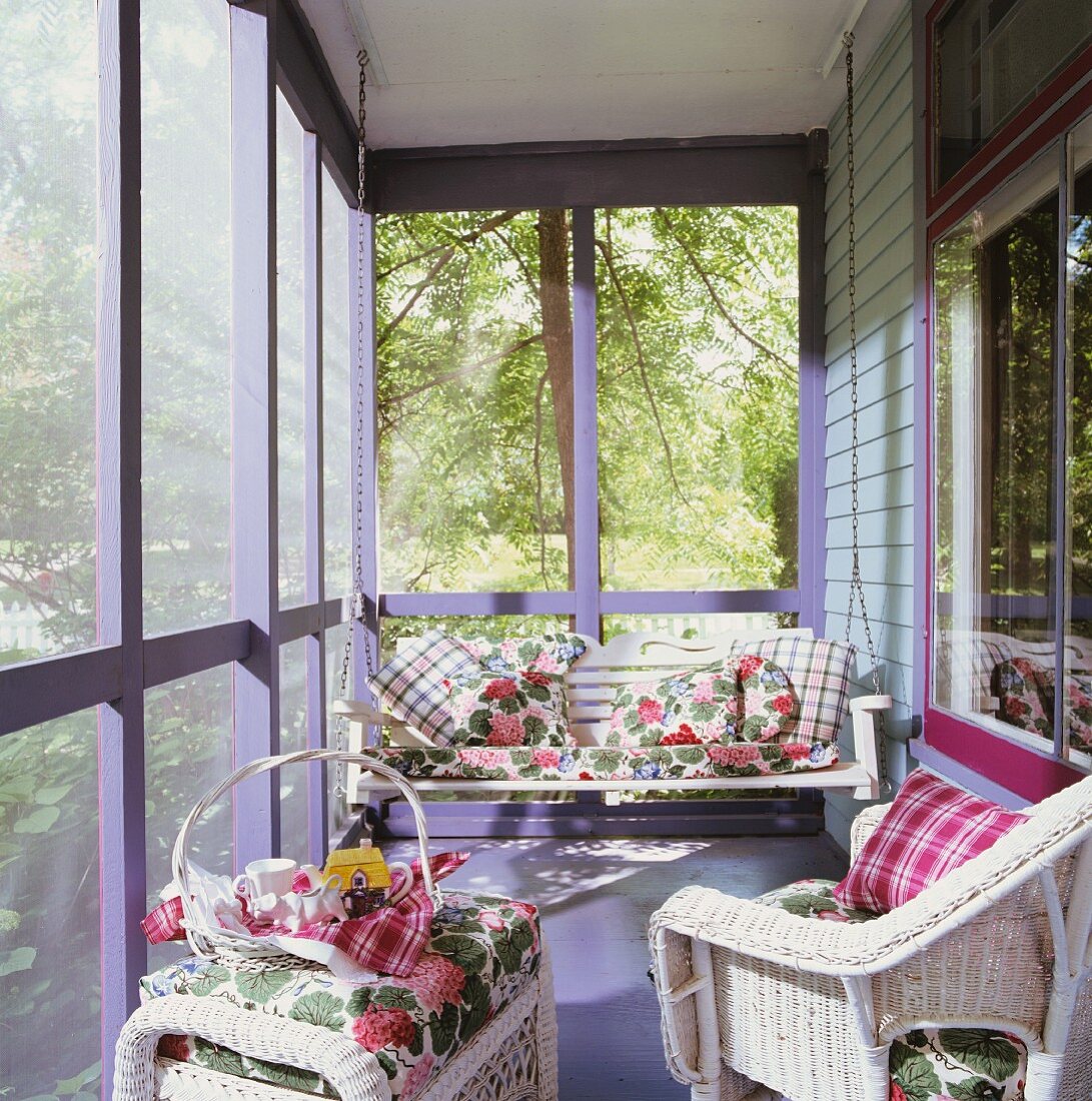 Verglaste, sonnige Veranda mit Hollywoodschaukel und Korbmöbeln; darauf Dekokissen mit Karo- und mit floralem Muster
