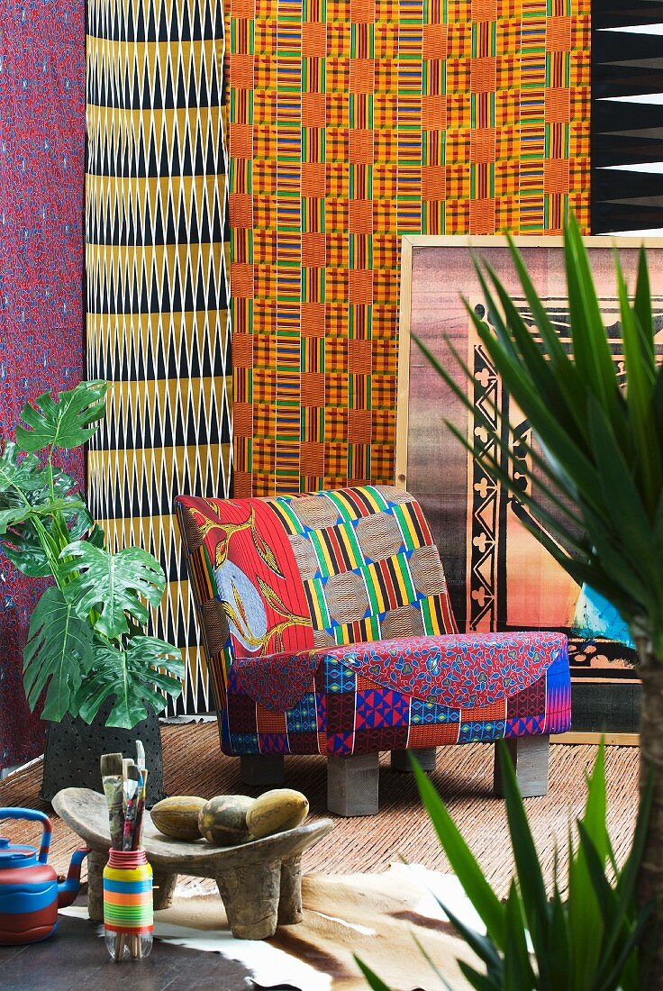 African style - colourful patterned chair in front of bright wall hangings surrounded by tropical potted plants