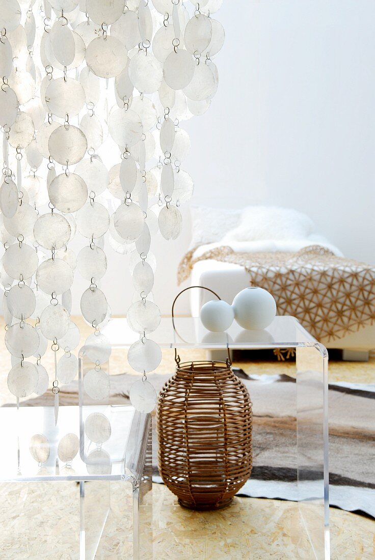 Effektvoller durchsichtiger Pailettenvorhang über Plexiglastischen und Korbvase, im Hintergrund Bett mit schlichter Tagesdecke