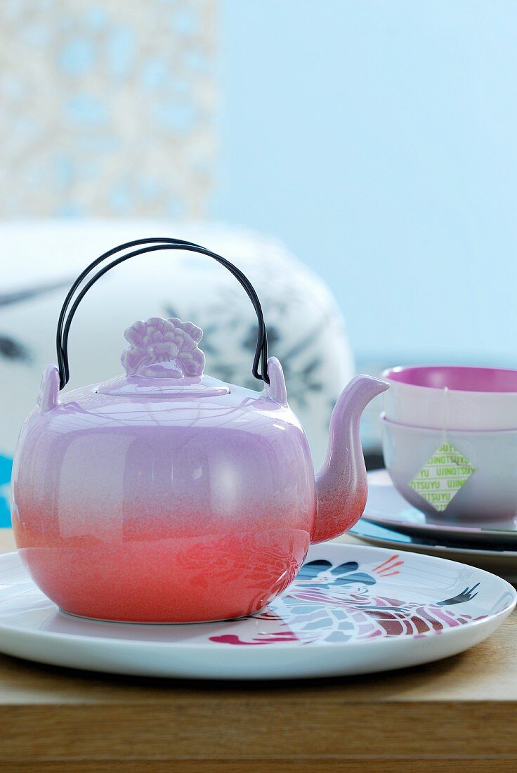 A tea set with a colourful teapot on a plate in front of a pale blue background