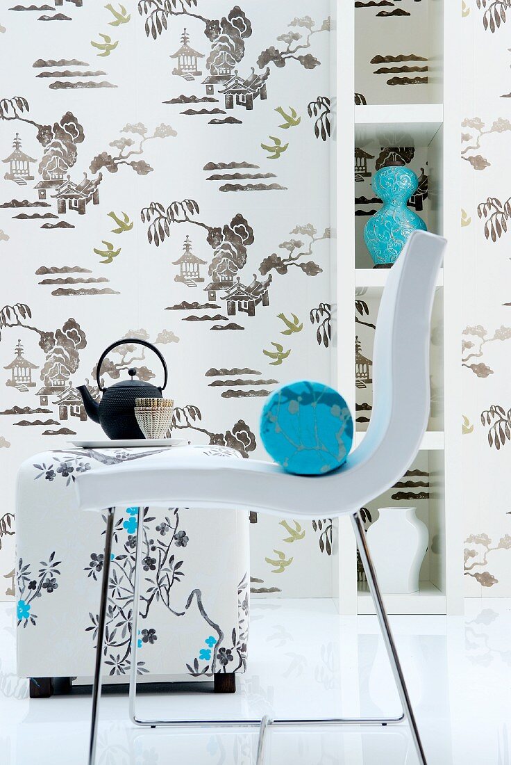 Moderner weißer Stuhl mit hellblauer Nackenrolle, dahinter gepolsterter Sitzhocker mit Teegeschirr vor tapezierter Wand und Regal mit Vasen