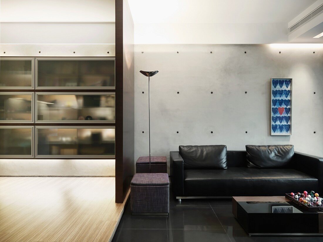 Offener Wohnraum mit schwarzer Ledercouch vor Betonwand neben Raumteiler mit Blick auf transparentem Einbauschrank