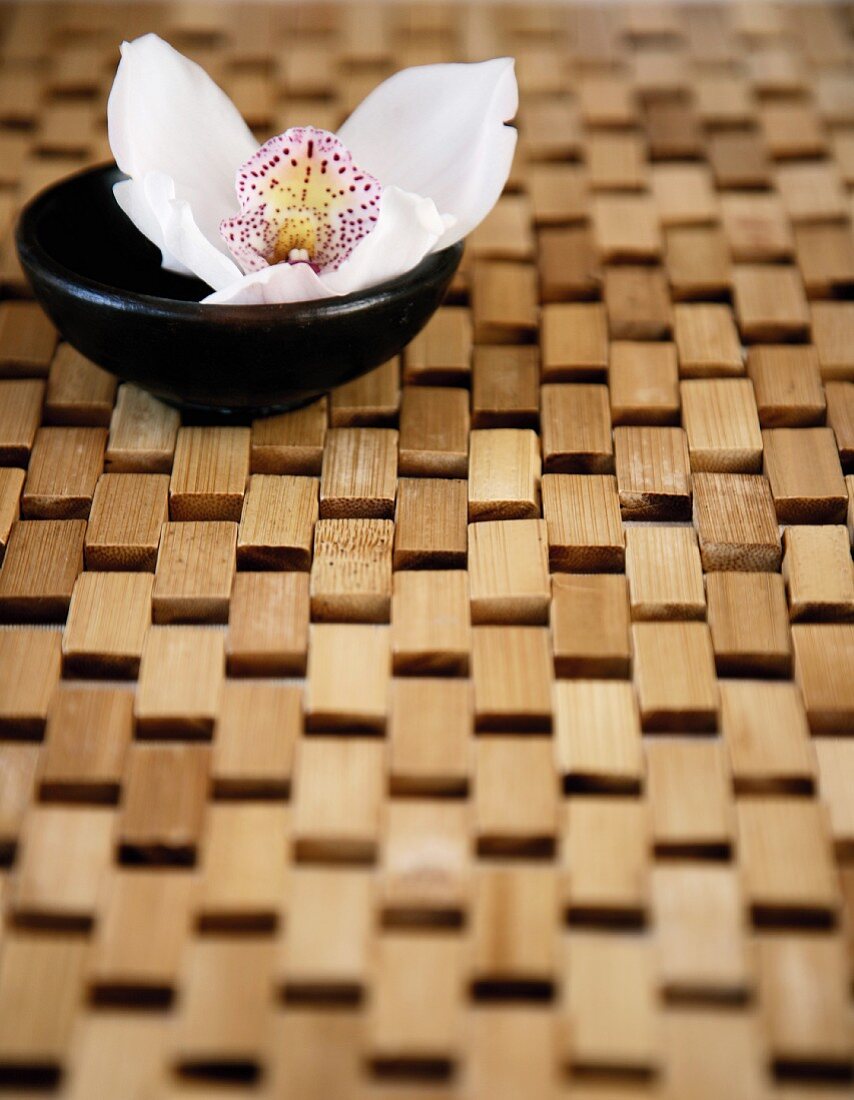 An orchid flower in a little black bowl on a bamboo-wood mat