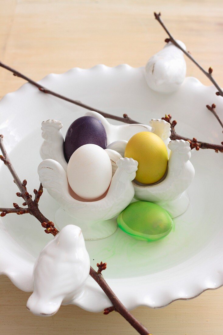 Bird ornaments, budding twigs and colourful Easter eggs in traditional egg cups arranged on white, china plate