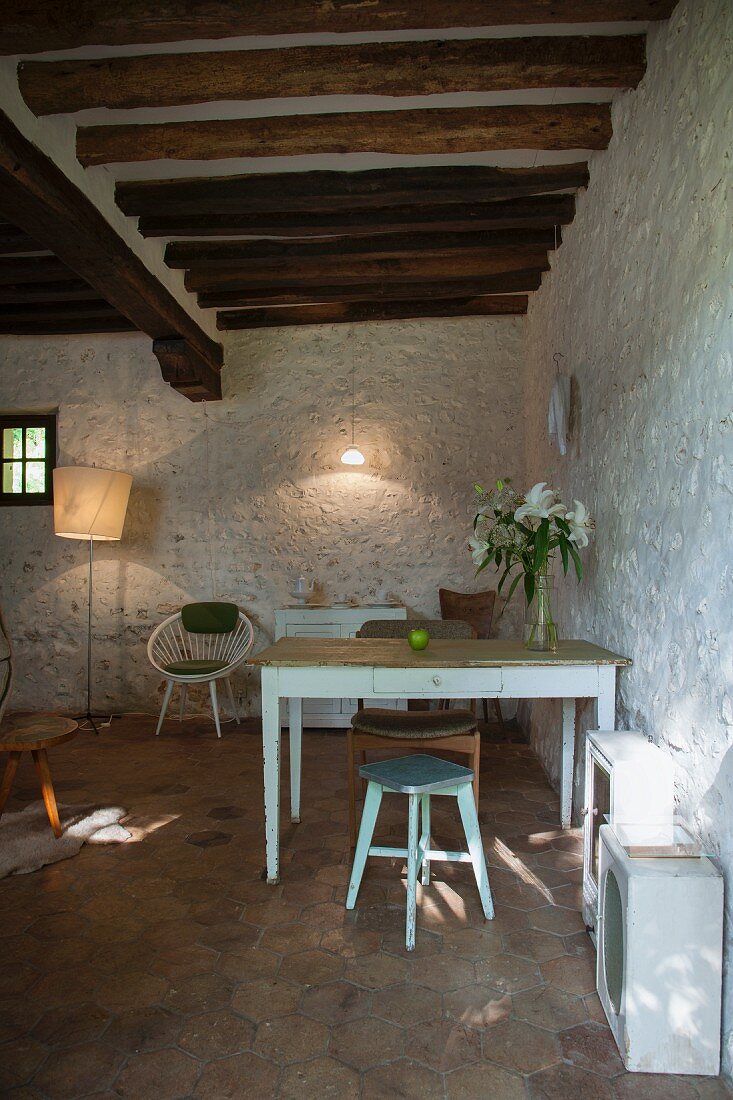 Kitchen table and wooden stools against white-painted stone wall below rustic wood-beamed ceiling