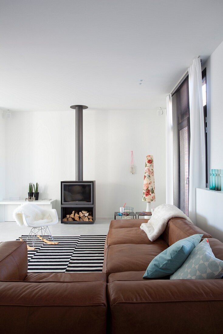 Braune Eckledercouch in minimalistischem Wohnraum, im Hintergrund vor Kaminofen Klassiker Schaukelstuhl mit Fell