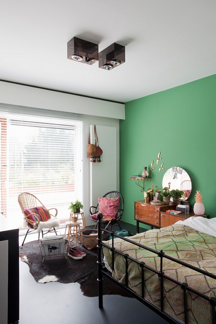 Black metal bed against green wall and wicker chair next to window in retro-style bedroom