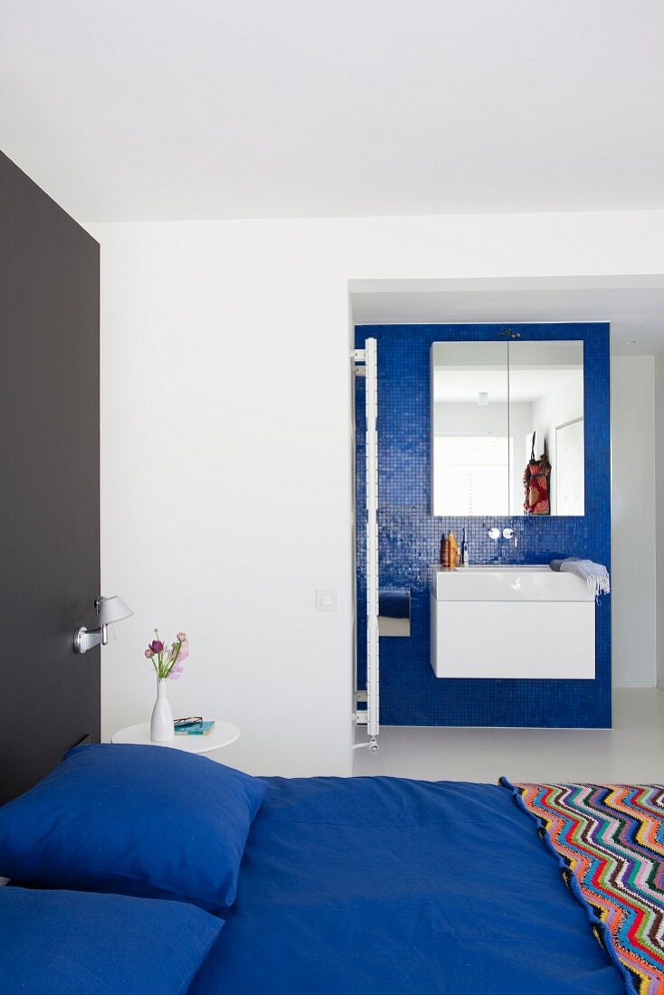 Königsblaue Bettwäsche auf Doppelbett vor schwarzer Wand und weisses Waschtischmöbel an blauer Mosaikfliesenwand in Bad Ensuite