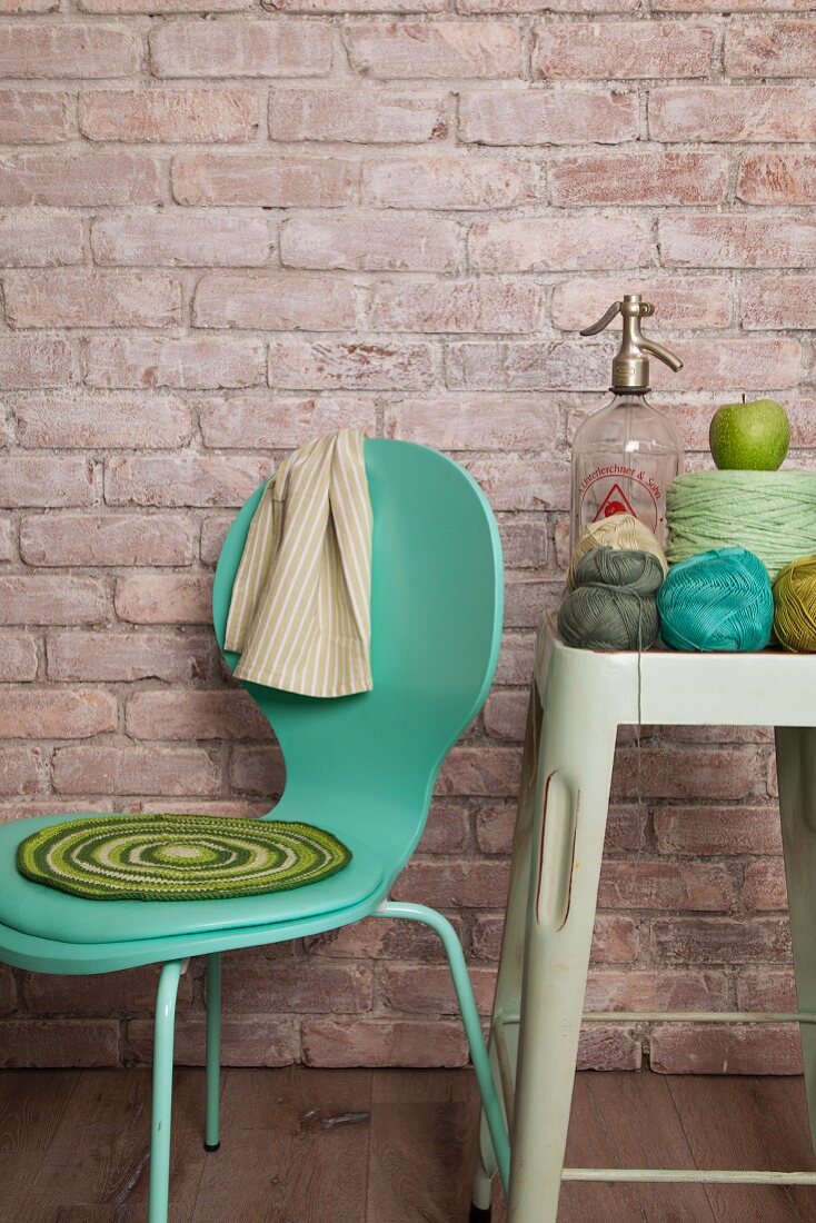 Gehäkelte Sitzauflage auf türkisgrünem Stuhl neben Häkelgarn und Sodaflasche auf Vintage Hocker vor Backsteinwand