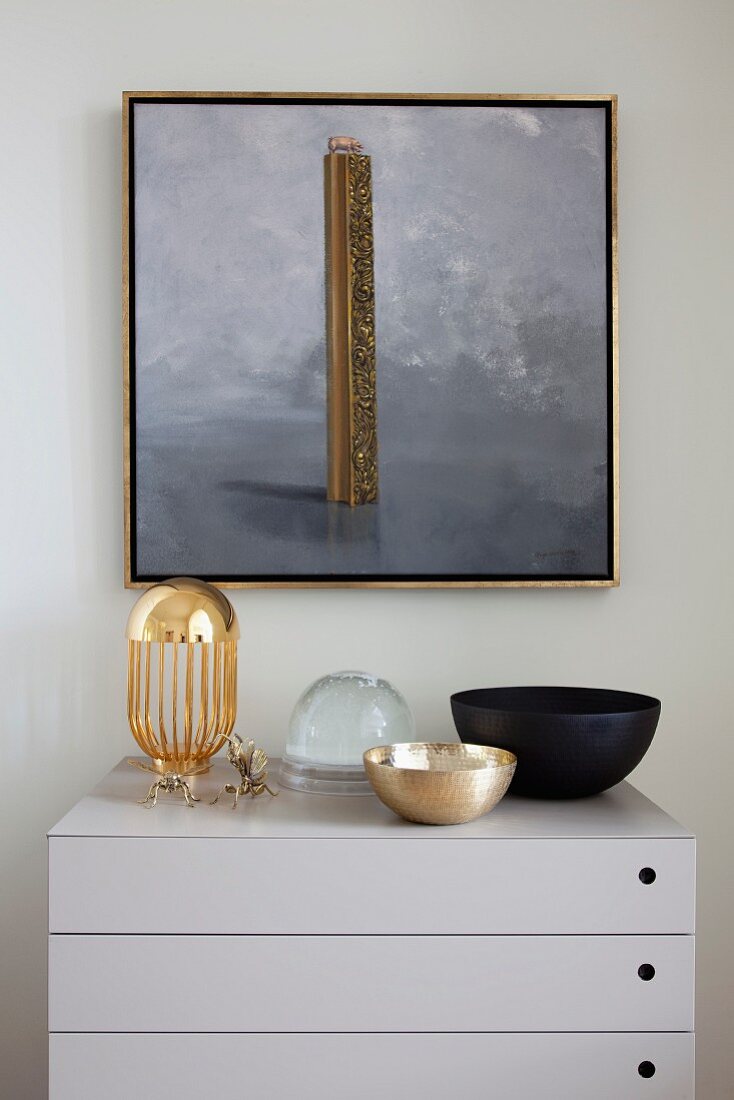Elegant bowls on modern, pale grey chest of drawers below gilt-framed picture on wall