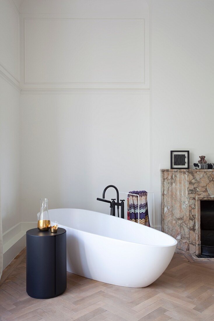 Free-standing white bathtub and cylindrical black side table on herringbone parquet floor in minimalist bathroom