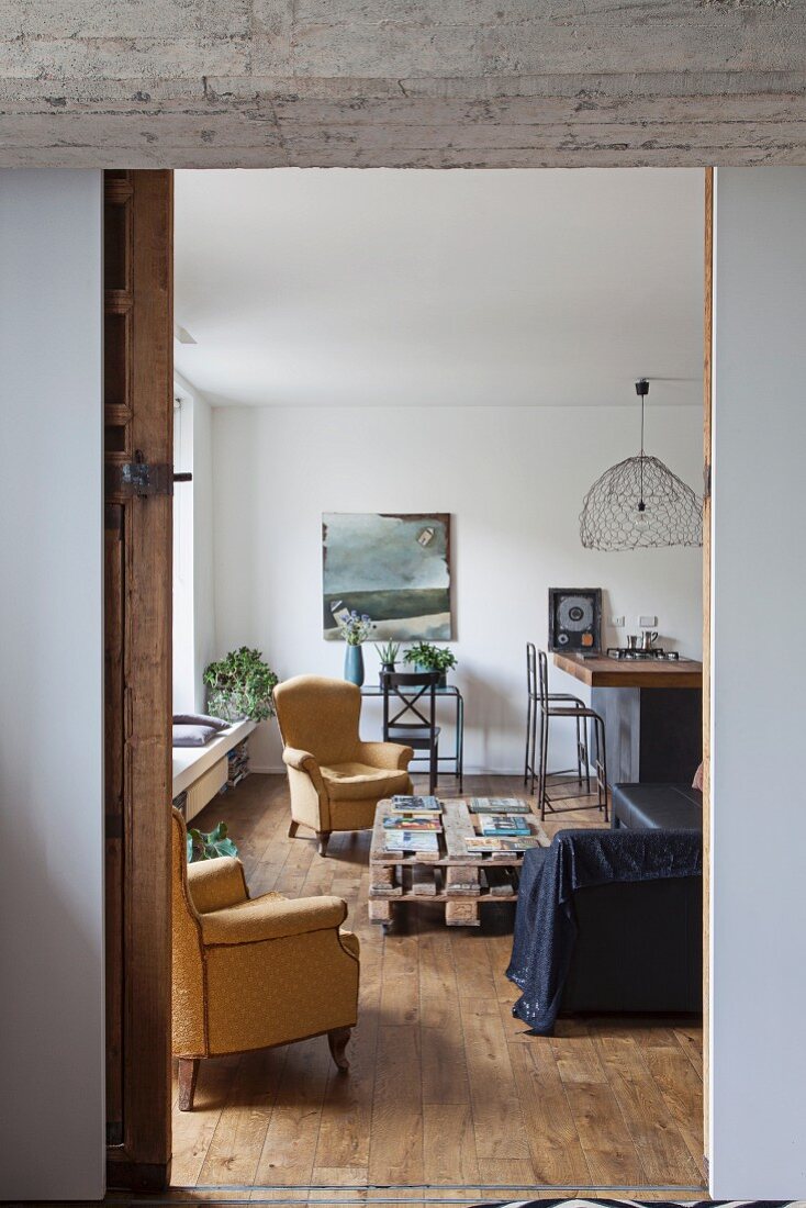 Blick auf Armlehnsessel um Paletten-Couchtisch in Wohnzimmer mit Küchentheke im Hintergrund