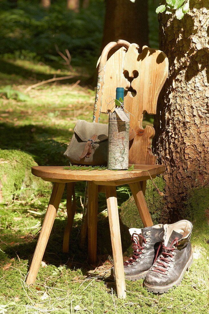 Holzstuhl vor Baum mit Wanderutensilien, Weinflasche mit Landkarte umwickelt und Wanderschuhe