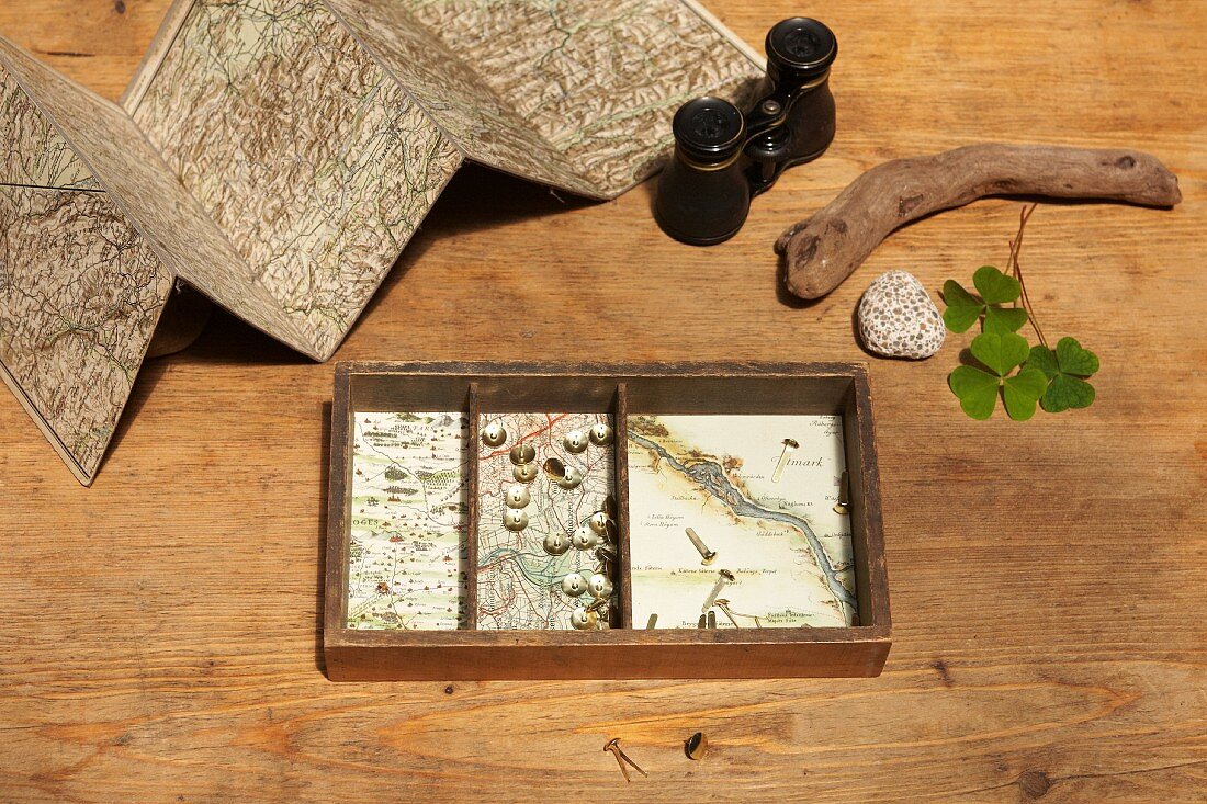 Old box lined with maps, binoculars, unfolded map and natural finds on wooden table