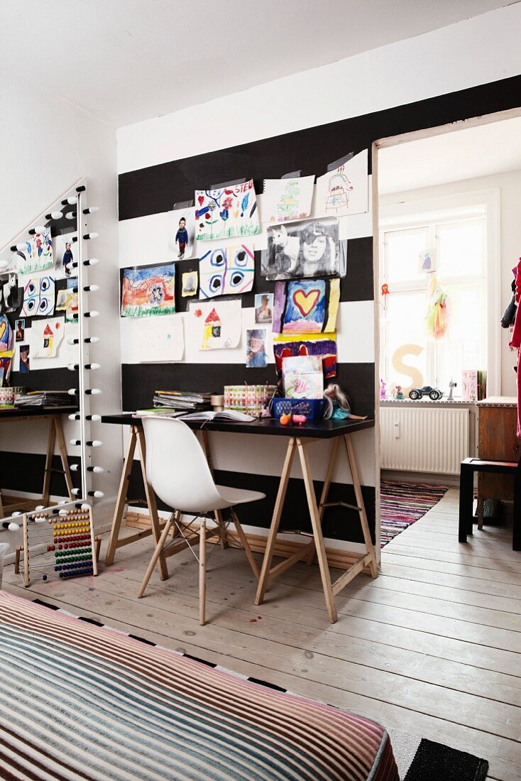Weißer Klassikerstuhl an Schreibtisch vor aufgehängten Kinderzeichnungen an schwarz-weiß gestreifter Wand mit Durchgang