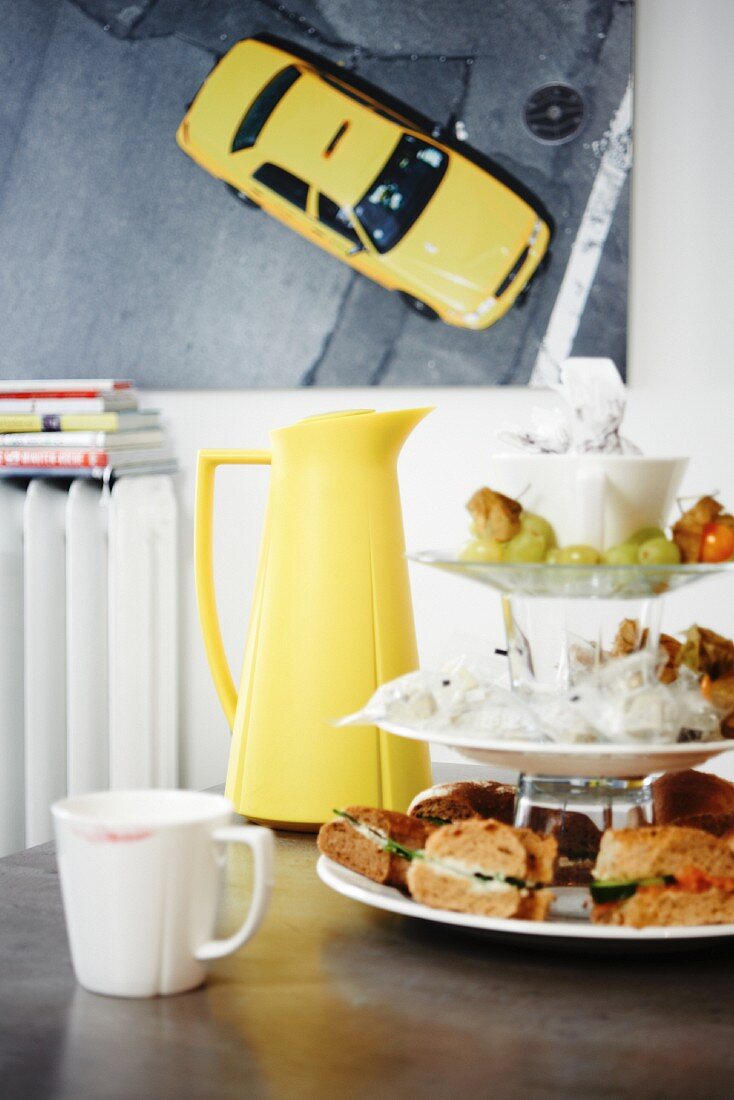 Cake stand, white coffee cup and yellow thermos flask in front of retro picture of car