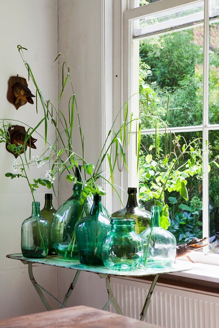Collection of demijohns in various shades of green, some holding leaves, on vintage folding table next to lattice window