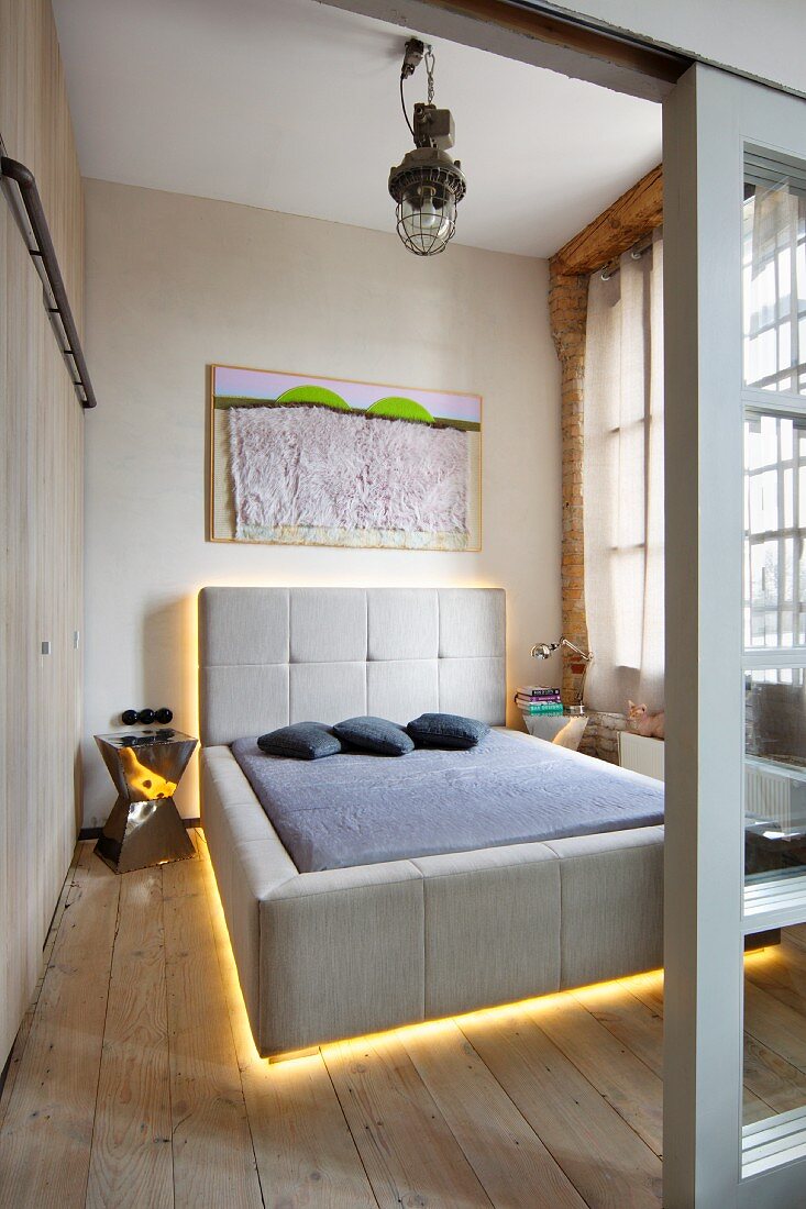 Blick durch offene Schiebetür auf hinterleuchtetes Designer-Doppelbett mit Polsterkopfteil und grauem Bezug in loftartigem Ambiente