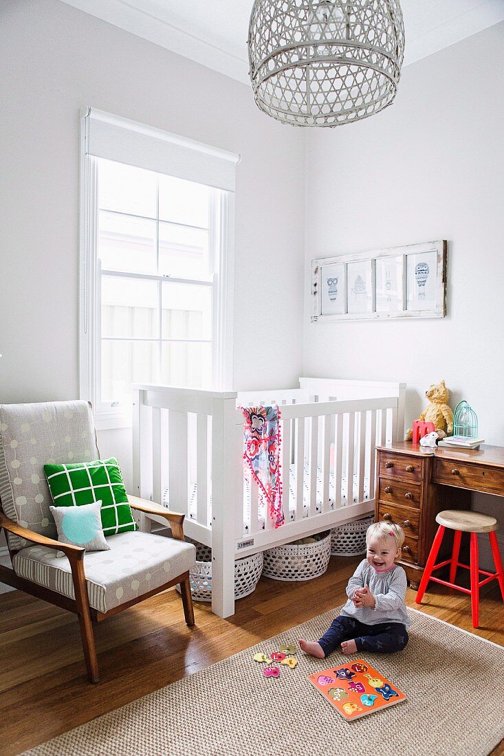 Toddler on sisal rug in front of white crib and retro armchair next to window in corner of room