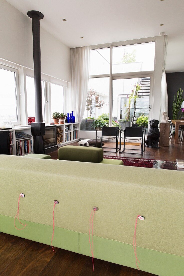 Rückseite eines Designersofas in Lindgrün in offenem Wohnraum, im Hintergrund Stühle vor Glasfront