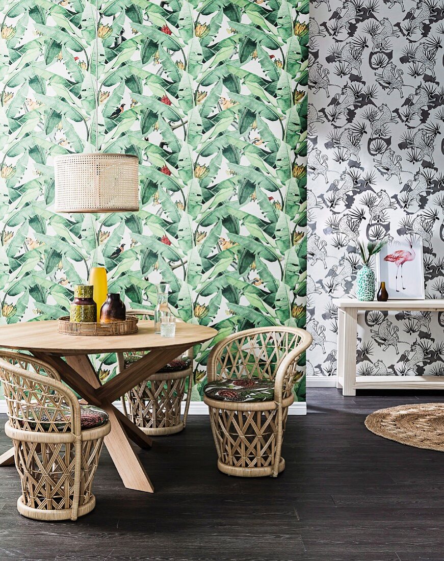 Round dining table with small wicker armchairs in front of wallpaper with green leaf motif, behind it is a black and white patterned wallpaper
