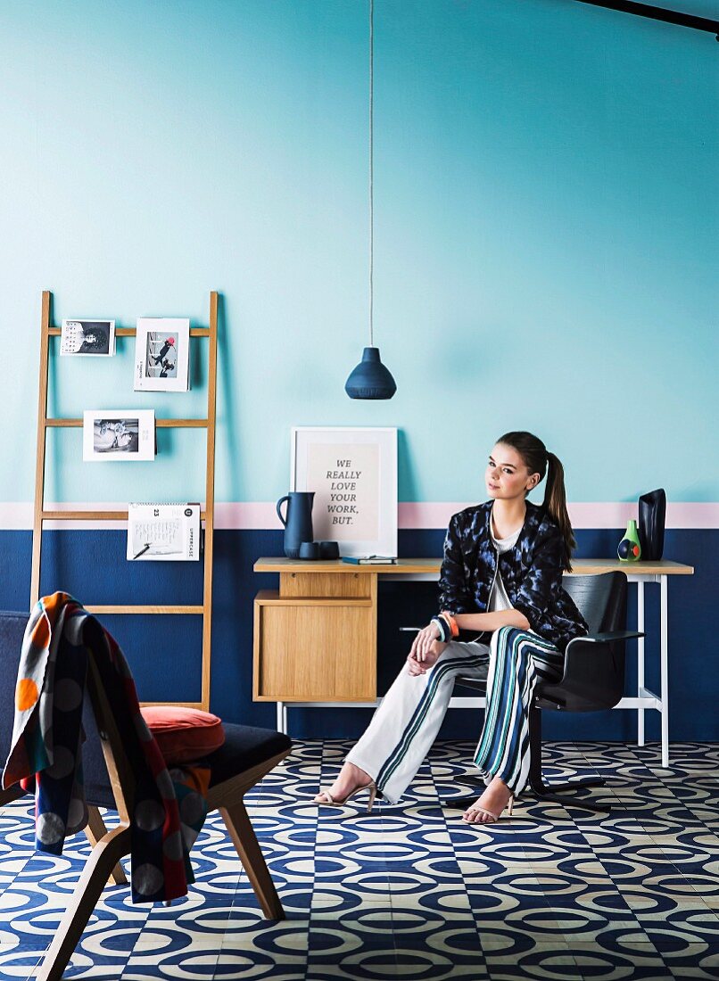 Frau auf Drehsessel an Schreibtisch und Dekoleiter vor blauem Wandsockel, dekorativer Boden mit Kreismuster, vorne ein skandinavischer Polsterstuhl