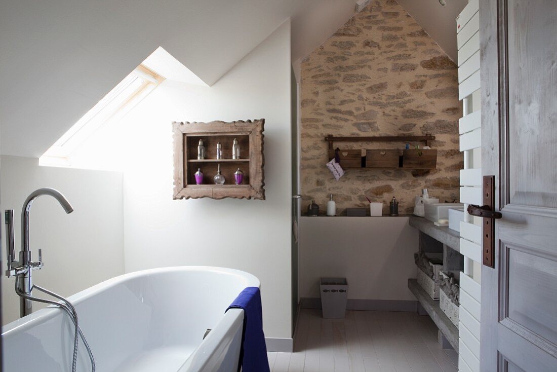 Blick durch offene Tür auf weiße Badewanne unter Dachfenster und Dachschräge, im Hintergrund Natursteinwand