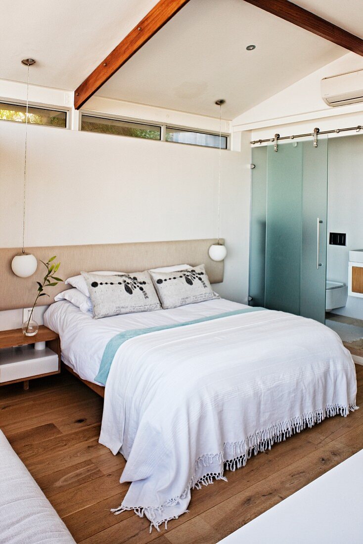 Doppelbett mit weisser Tagesdecke vor Wand mit Oberlicht Schlitzen, seitlich Bad ensuite mit halboffener Glas Schiebetür, in modernem Schlafzimmer