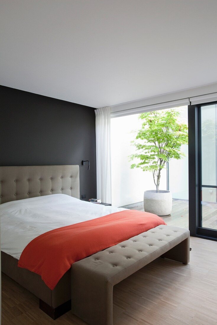Elegantes Doppelbett an schwarzer Wand mit taupefarbenem Kopfteil und passender Kleiderbank, Blick durch offene Schiebetür in Patio mit Kübelpflanze