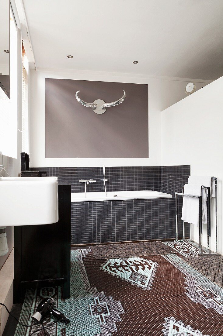 Modernes Bad mit gemustertem Teppich, im Hintergrund Badewanne mit schwarzen Fliesen, oberhalb stilisierte Tiertrophäe