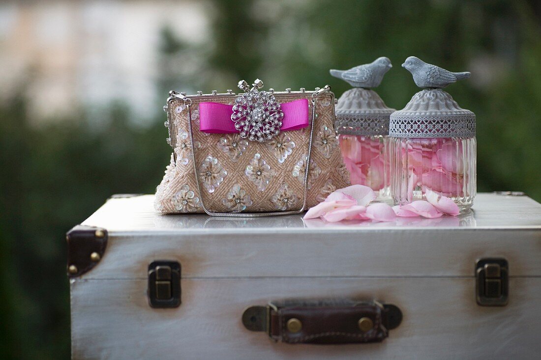 Edles Paillettentäschchen mit Brosche und pinkfarbenem Schleifenband neben Glasgefäßen mit Rosenblütenblättern und Vogelfiguren auf Vintage Koffer