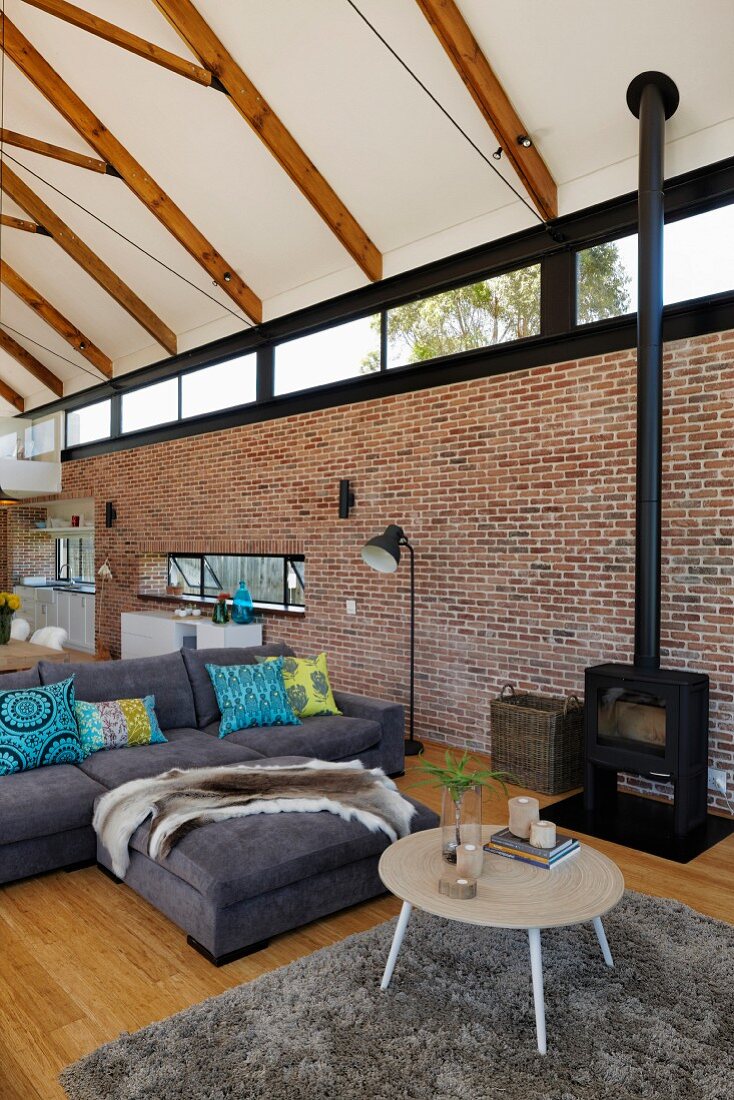 Coffee table, flokati rug and grey sofa in lounge area of open-plan interior with exposed roof structure and brick wall