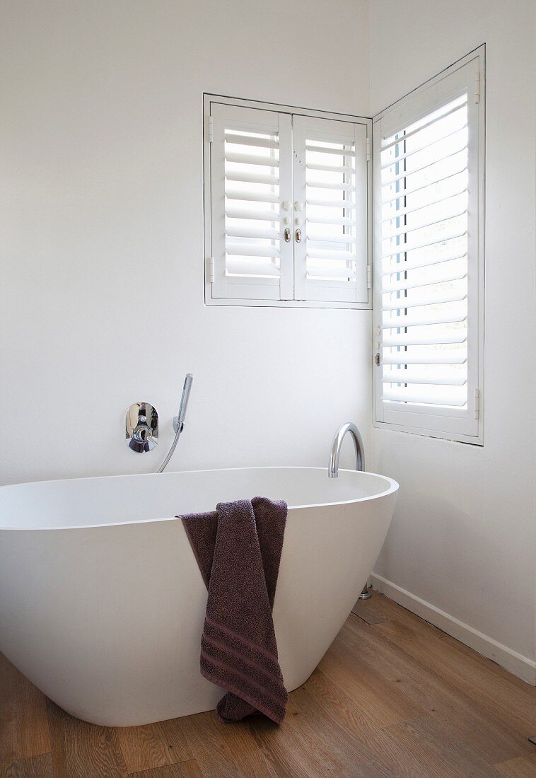 weiße freistehende Designer-Badewanne mit dunkelbraunem Handtuch auf Eichenparkett vor Übereckfenster mit Fensterläden