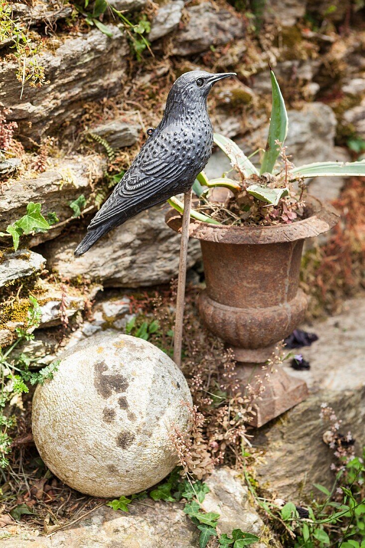 Vogelfigur auf Holzstab neben antikem Pflanzgefäss und Steinkugel gesteckt