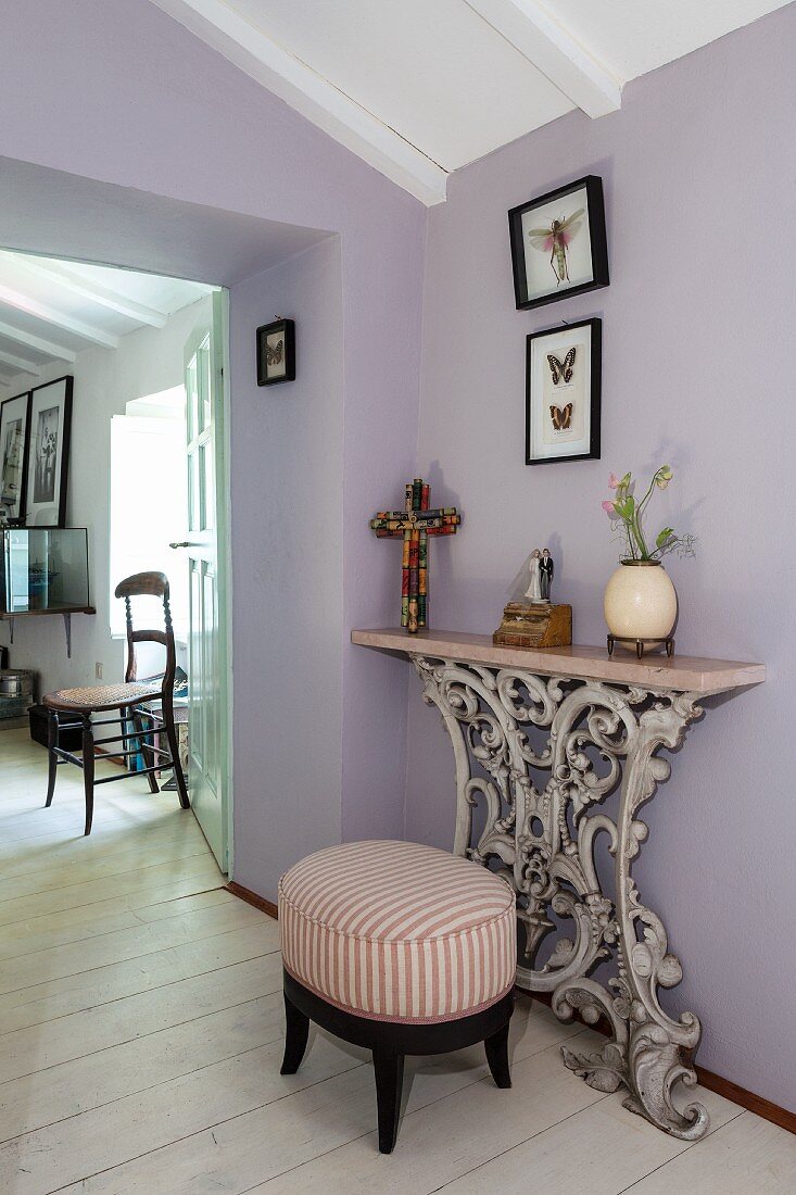 Antique stool with striped upholstery next to console table with ornamental base against lilac wall