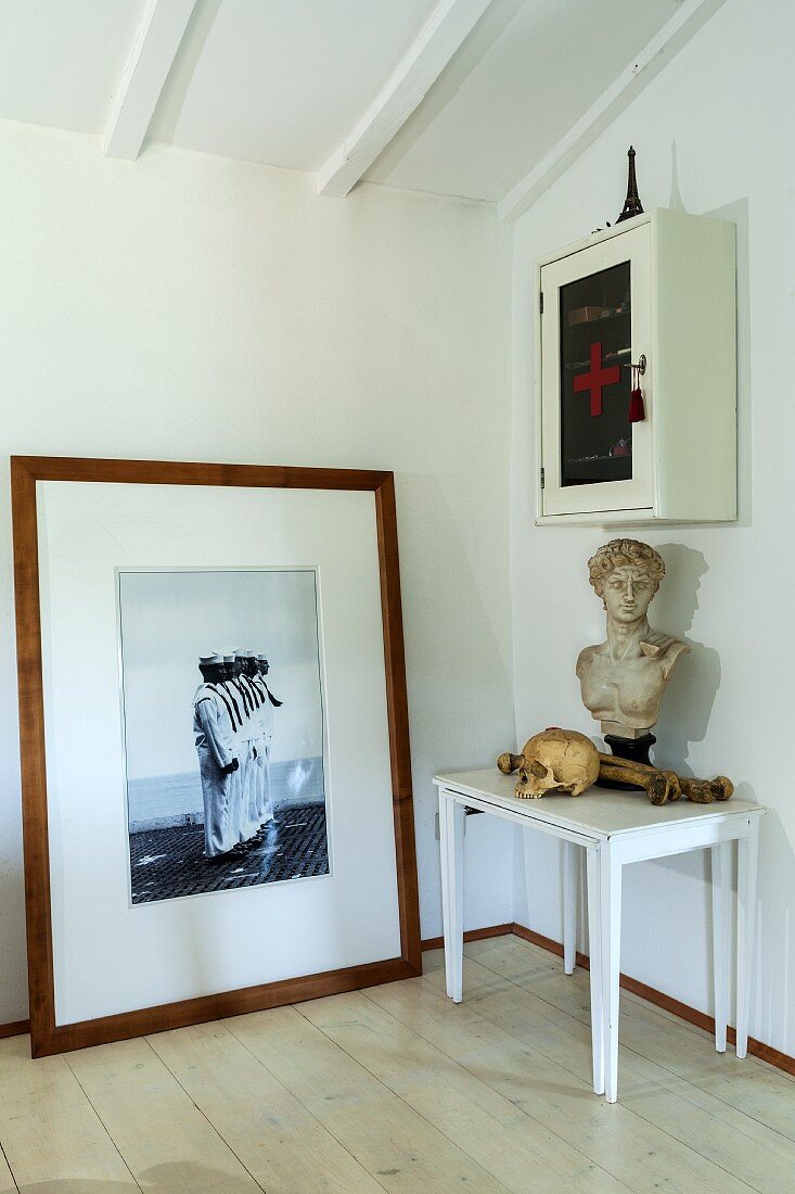 Framed photo leaning against wall, bust and skull on set of white side tables and glass-fronted cabinet on wall