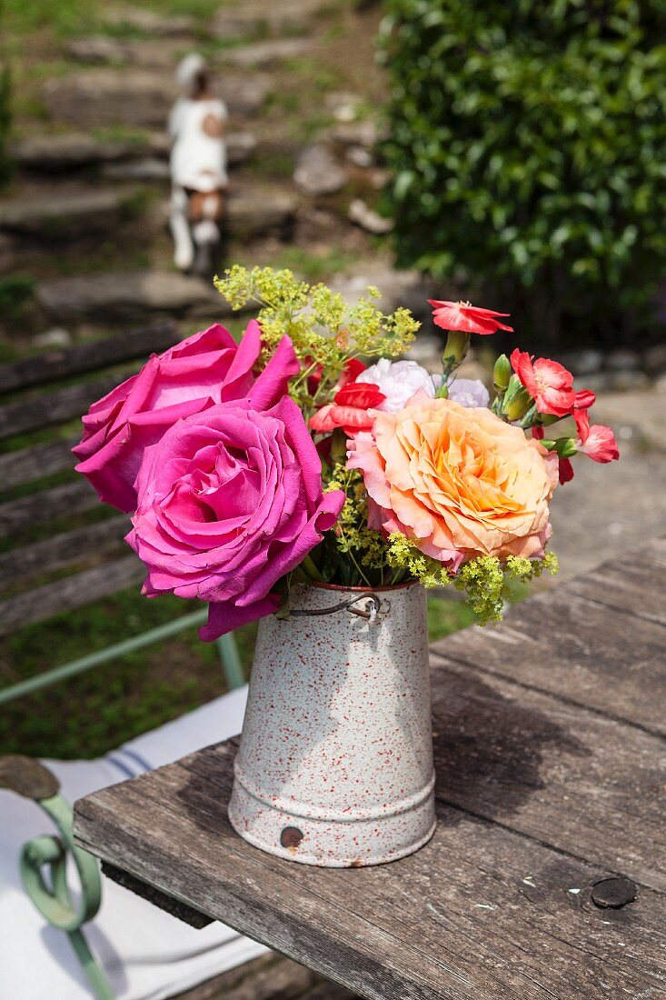 Blumenstrauss mit Rosen und Nelken in Vintage Kanne auf rustikalem Gartentisch