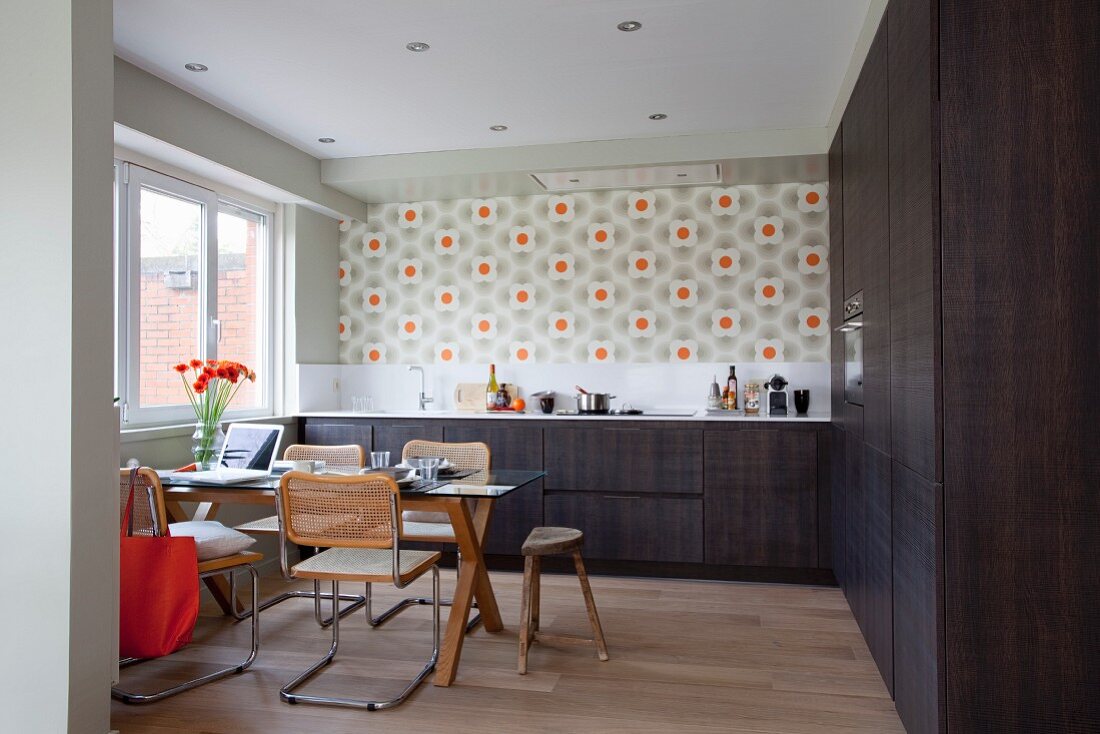 Cantilever chairs around dining table in kitchen with dark brown cabinets and retro floral wallpaper