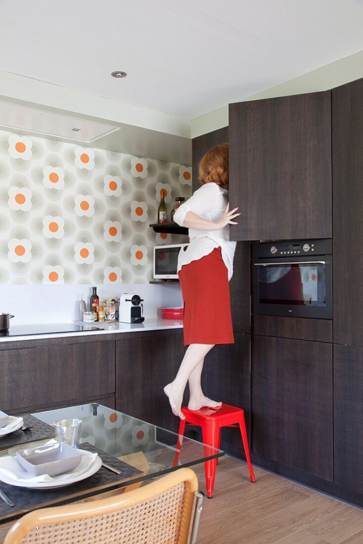 Woman standing on red stool in front of fitted kitchen cupboards with dark brown wooden doors