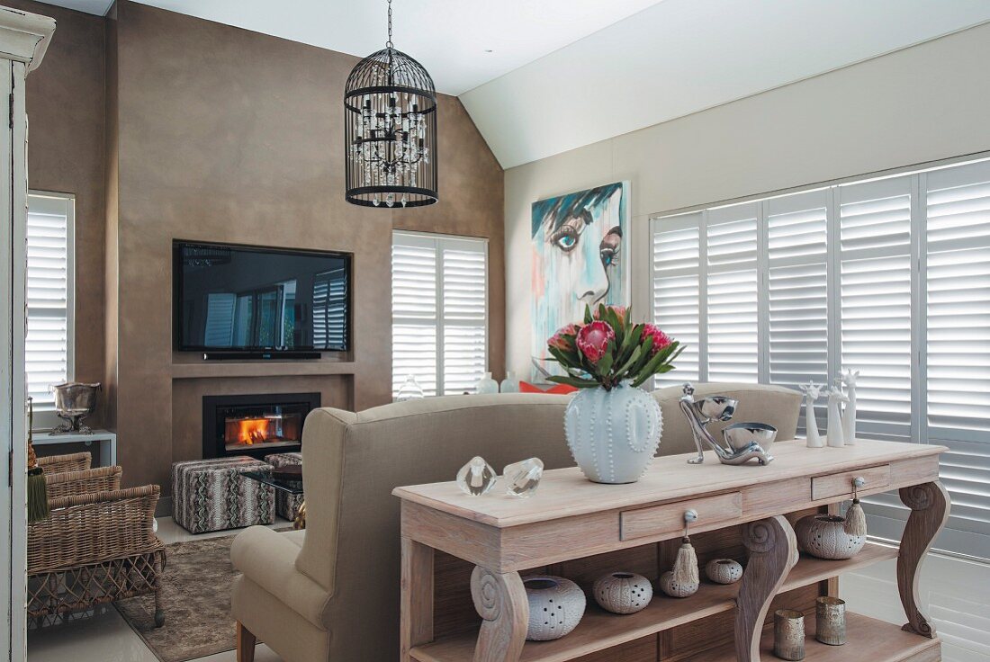 Loungebereich, Konsolentisch aus Holz mit Schnitzereien vor Rückseite eines Sofas, gegenüber Flachbildschirm an hellbraun getönter Wand
